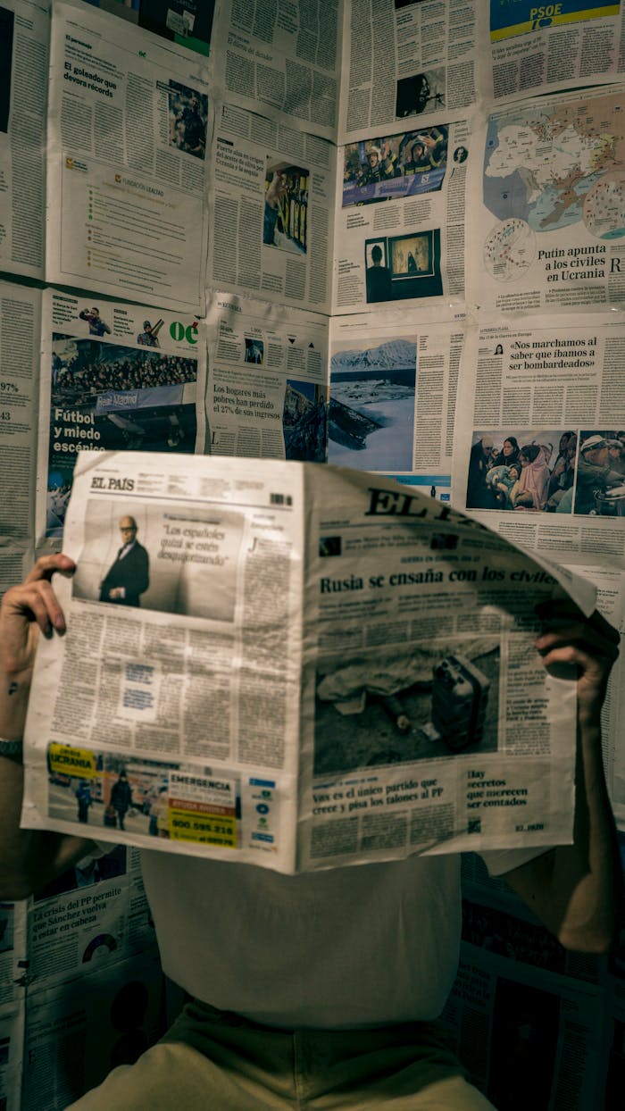A person reads a newspaper in a room with walls covered in news articles.